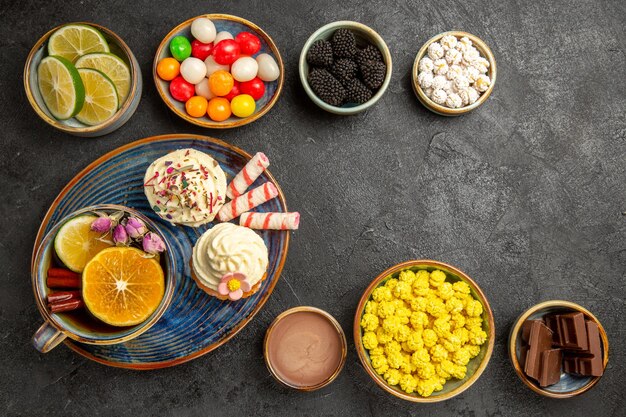 Vista dall'alto dolci sul tavolo ciotole di frutti di bosco cioccolato agrumi dolci colorati e un piatto di cupcakes e una tazza di tisana con bastoncini di cannella sul tavolo scuro