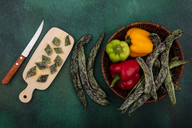 Vista dall'alto fagiolini con peperoni in un cesto con un tagliere con un coltello su uno sfondo verde