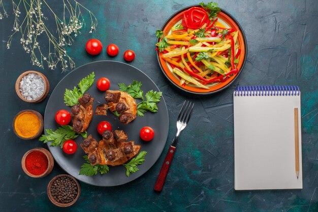 Vista dall'alto fette di carne fritta con blocco note di insalata di verdure a fette e condimenti sul pasto salutare di carne di cibo vegetale da scrivania blu scuro