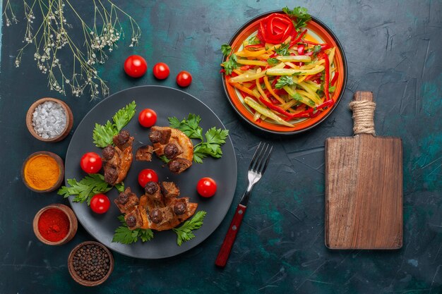 Vista dall'alto fette di carne fritta con insalata di verdure a fette e condimenti sul pasto salutare di carne di cibo di verdure da scrivania blu scuro