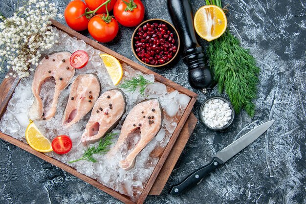 Vista dall'alto fette di pesce crudo con ghiaccio su ciotole di tavola di legno con semi di melograno sale marino aneto pomodori coltello su sfondo grigio