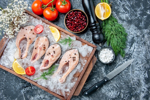 Vista dall'alto fette di pesce crudo con ghiaccio su ciotole di tavola di legno con semi di melograno sale marino aneto pomodori coltello su sfondo grigio