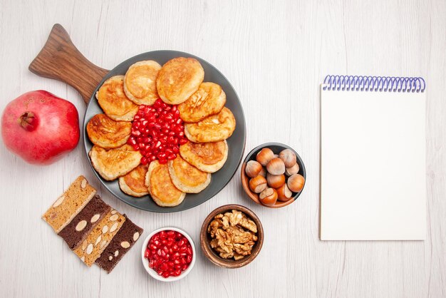 Vista dall'alto frittelle e melograno al melograno accanto al quaderno bianco ciotole di marmellata e nocciole piatto da torta di frittelle e semi di melograno sul tagliere di legno sul tavolo