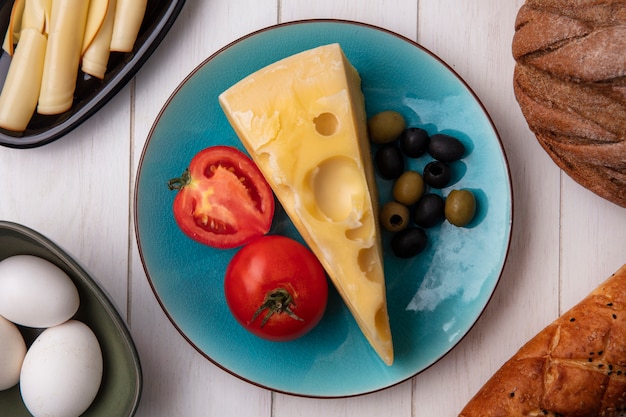 Vista dall'alto il formaggio maasdam con pomodori e olive su un piatto con uova di gallina e una pagnotta di pane bianco e nero su una piastra bianca