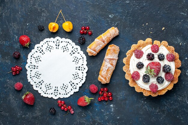 Vista dall'alto in lontananza deliziosa piccola torta con crema e frutti di bosco insieme a braccialetti sulla scrivania scura