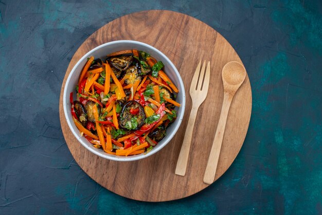 Vista dall'alto insalata di verdure a fette all'interno del piatto con posate di legno sulla superficie blu scuro