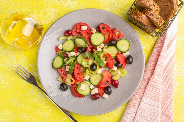 Vista dall'alto insalata di verdure fresche con cetrioli affettati pomodori oliva all'interno della piastra con olio e pane sul colore del pasto di insalata di cibo vegetale superficie gialla