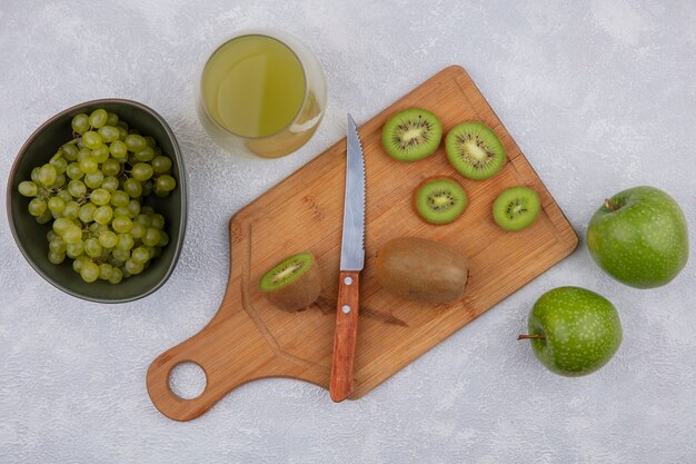 Vista dall'alto le fette di kiwi con un coltello su un tagliere con mele verdi e uva in una ciotola con succo di mela in un bicchiere su sfondo bianco