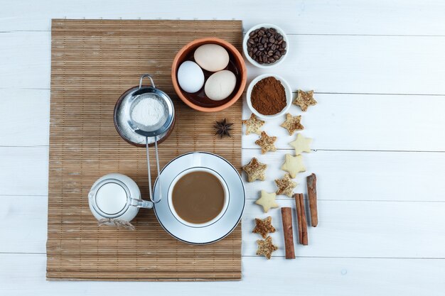 Vista dall'alto le uova, la brocca di latte, la tazza di caffè, la farina sulla tovaglietta con i chicchi di caffè e la farina, i biscotti della stella, la cannella sul fondo bianco del bordo di legno. orizzontale