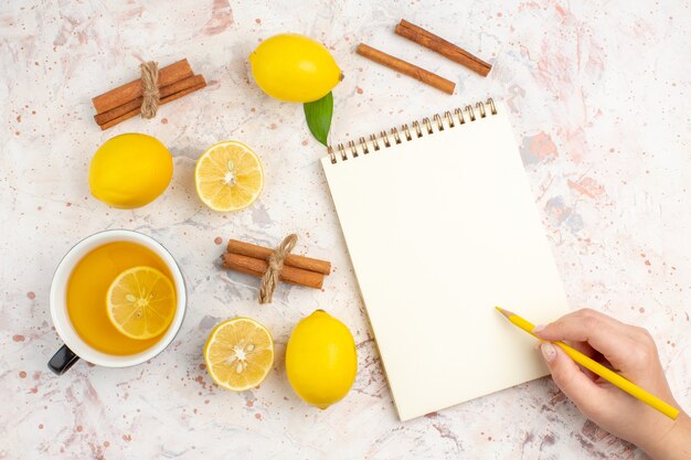 Vista dall'alto limoni freschi tagliati bastoncini di cannella al limone una tazza di tè al limone matita in un blocco note di mano femminile sulla superficie isolata luminosa