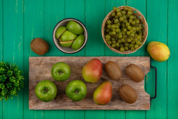 Vista dall'alto mele verdi con kiwi e pera su un tagliere e uva verde in un cesto su uno sfondo verde