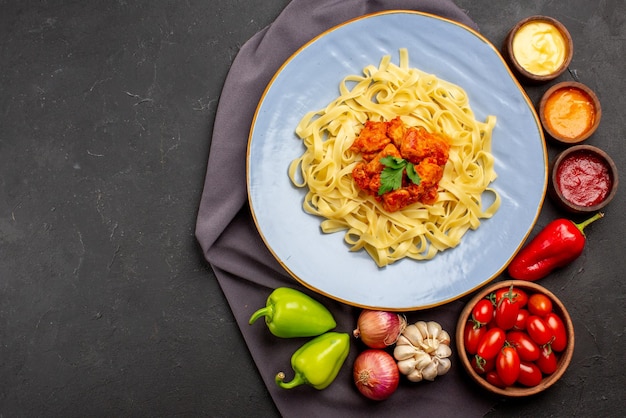 Vista dall'alto pasta su tovaglia piatto di pasta appetitosa accanto alla ciotola di pomodori e salse colorate aglio cipolla peperone sulla tovaglia viola sul tavolo
