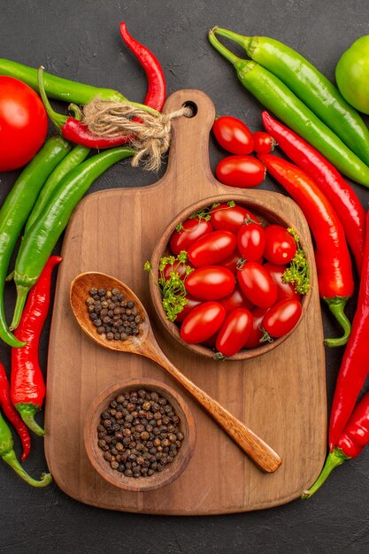 Vista dall'alto peperoni rossi e verdi caldi pomodori ciotole con pomodorini e pepe nero e cucchiaio su un tagliere su fondo nero