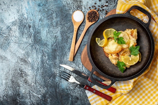 Vista dall'alto pesce fritto in padella con limone e prezzemolo diverse spezie in cucchiai di legno forchetta e coltello su sfondo grigio