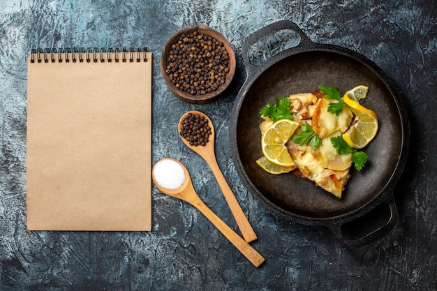 Vista dall'alto pesce fritto in padella con limone e prezzemolo spezie in cucchiai di legno pepe nero in una ciotola notebook su sfondo grigio