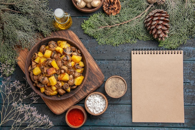 Vista dall'alto piatto di ciotola di funghi e patate sul tagliere accanto a diverse spezie e taccuino sotto una bottiglia di olio ciotola di funghi bianchi e rami di abete con coni