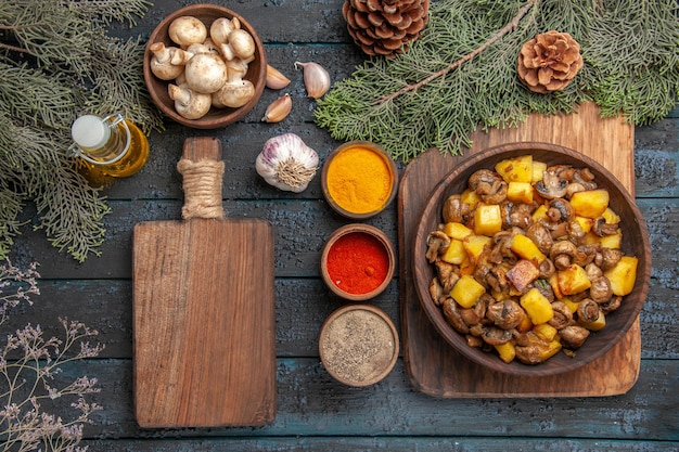 Vista dall'alto piatto e piatto di funghi e patate su tavola di legno accanto a spezie colorate tagliere olio in bottiglia aglio ciotola di funghi e rami con coni