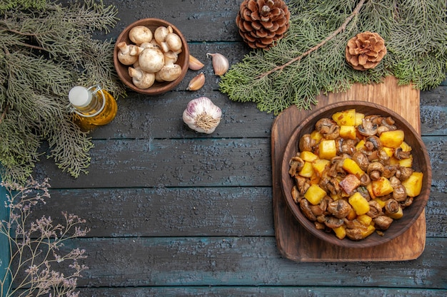 Vista dall'alto piatto e rami piatto di funghi con patate sul tagliere accanto all'olio in bottiglia aglio ciotola di funghi e rami con coni