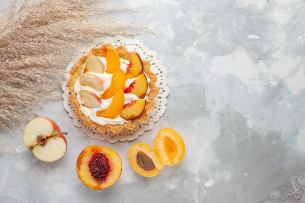 Vista dall'alto piccola torta cremosa con frutta a fette e crema bianca insieme a pesche albicocche fresche sulla scrivania a luce bianca, torta di frutta, biscotto biscotto