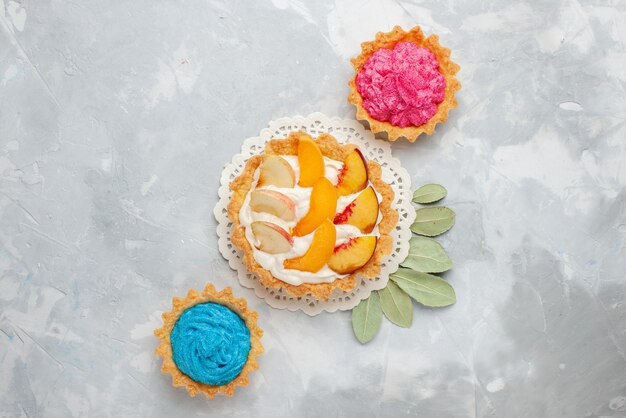 Vista dall'alto piccola torta cremosa con frutta a fette e crema bianca insieme a torte cremose sulla scrivania luce bianca torta di frutta biscotto biscotto dolce