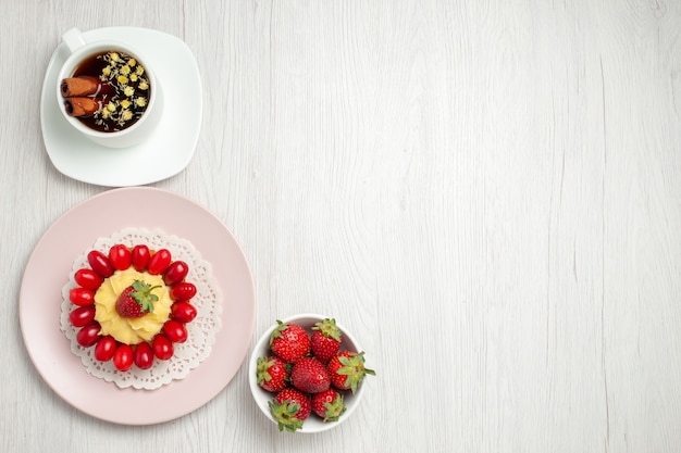 Vista dall'alto piccola torta cremosa con frutta e tazza di tè sulla scrivania bianca