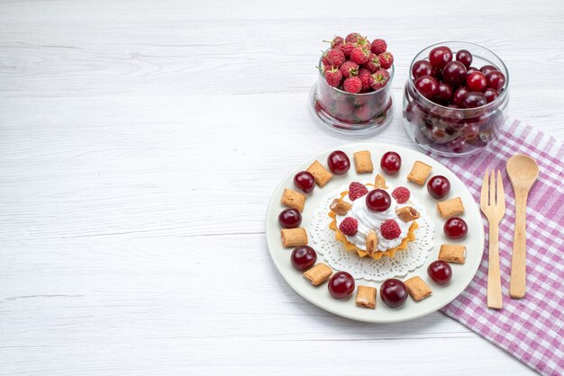 Vista dall'alto piccola torta cremosa con lamponi e piccoli biscotti insieme a ciliegie sulla scrivania a luce bianca torta alla frutta dolce crema di bacche ciliegia