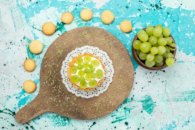 Vista dall'alto piccola torta gustosa con crema deliziosa e biscotti di uva fresca e affettata sulla frutta zucchero dolce torta da scrivania luce blu