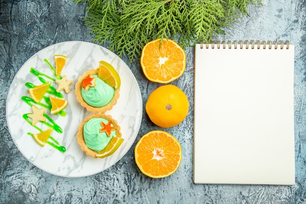 Vista dall'alto piccole crostate con crema pasticcera verde e fetta di limone sul blocco note di arance tagliate piatto sul tavolo scuro