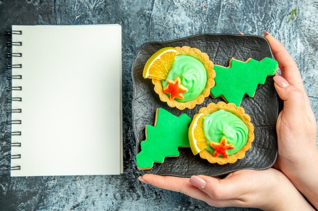 Vista dall'alto piccole torte biscotti dell'albero di Natale sulla banda nera in mano femminile notebook sul tavolo grigio