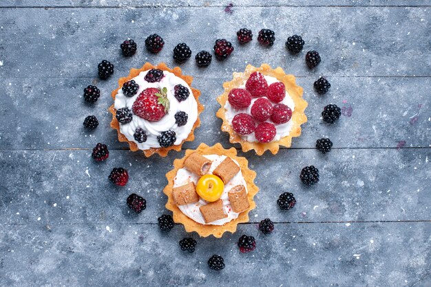 Vista dall'alto piccole torte cremose con lamponi insieme a more a forma di cuore sulla foto del biscotto torta di frutti di bosco luminoso scrivania