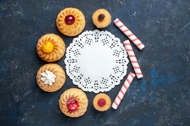 Vista dall'alto piccole torte deliziose insieme a caramelle rosa stick sul tavolo scuro torta di biscotti dolci cuocere