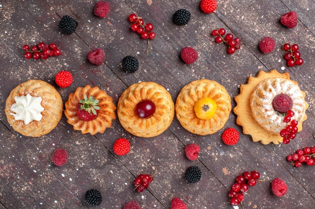 Vista dall'alto piccole torte e biscotti deliziosi con frutti di bosco diversi lungo tutto il biscotto della foto della bacca del biscotto della torta della scrivania marrone