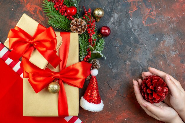 Vista dall'alto piccoli regali legati con nastro rosso cappello da Babbo Natale ramo di albero di Natale pigna in mani femminili sul tavolo rosso scuro