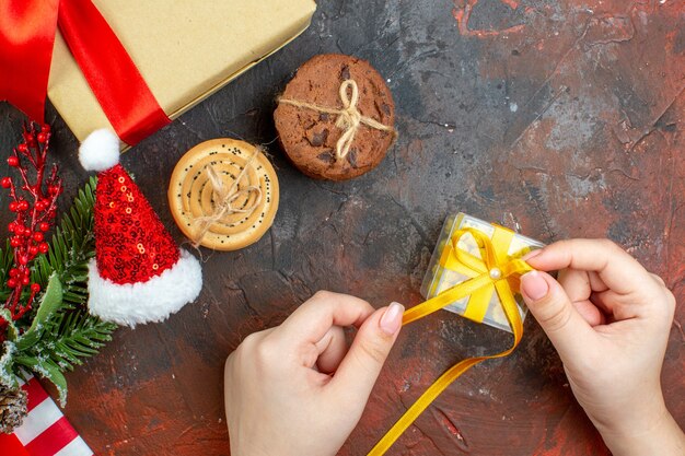 Vista dall'alto piccolo regalo in mano femminile biscotti con cappello di Babbo Natale sul tavolo rosso scuro