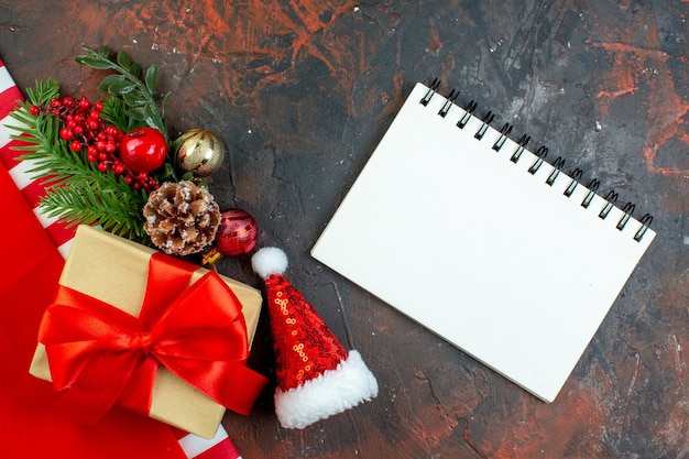 Vista dall'alto piccolo regalo legato con nastro rosso ramo di un albero di Natale mini blocco note cappello di Babbo Natale sul tavolo rosso scuro