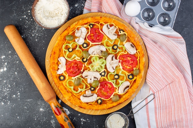 Vista dall'alto pizza ai funghi con pomodori rossi olive tutte tagliate all'interno con olio e farina sulla scrivania grigia impasto per pizza italiano