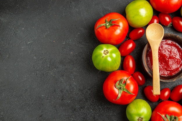 Vista dall'alto pomodori rossi e verdi ciliegia intorno a una ciotola con ketchup e un cucchiaio di legno sul tavolo scuro con spazio di copia