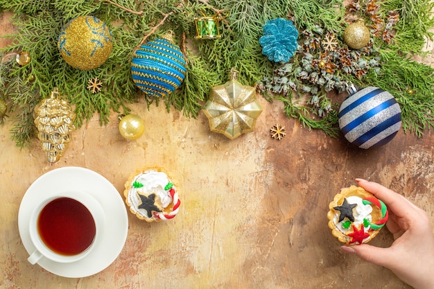 Vista dall'alto rami di abete albero di natale ornamenti una tazza di tè cupcake in mano femminile su sfondo beige