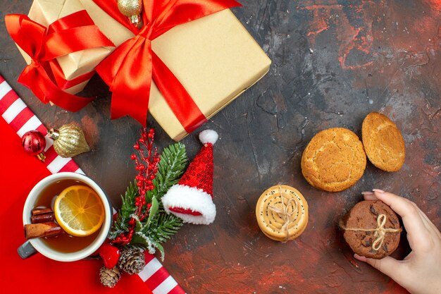 Vista dall'alto regali di Natale legati con nastro rosso biscotti cappello di Babbo Natale in mano femminile tazza di tè sul tavolo rosso scuro