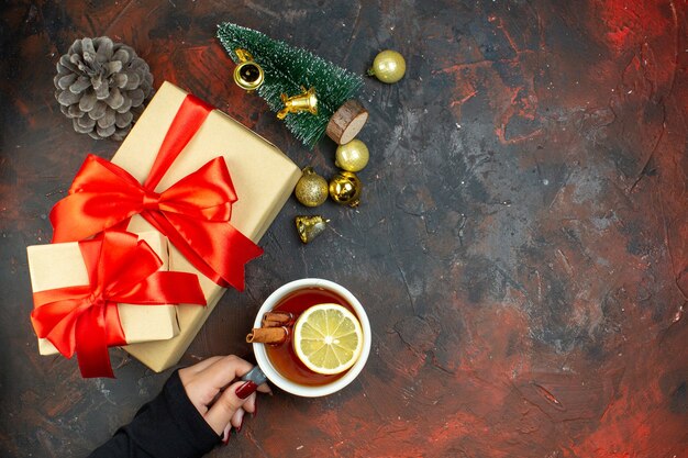 Vista dall'alto regali di natale palline di natale dorate tazza di tè in mano femminile mini albero di natale sul tavolo rosso scuro posto libero