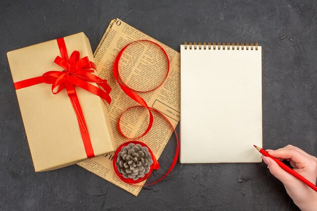 Vista dall'alto regalo di natale in nastro di carta marrone sul giornale un quaderno matita rossa in mano femminile su superficie scura