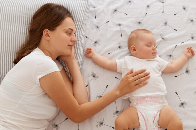 Vista dall'alto ritratto di donna che dorme con la sua piccola figlia sul letto, mamma sdraiata con bambina che giura maglietta bianca, maternità felice e infanzia.