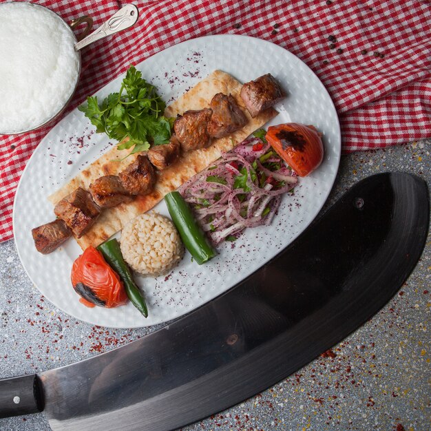 Vista dall'alto shish kebab con verdure fritte e cipolla tritata e ayran e coltello nel piatto bianco