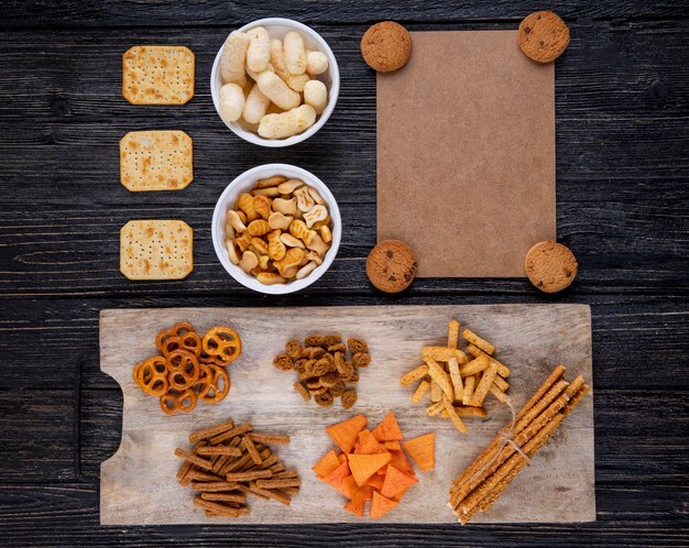 Vista dall'alto snack biscotti al cioccolato fette biscottate brezel paprika chip cracker bastoncini di pesce cracker e mais bastoni su fondo di legno nero