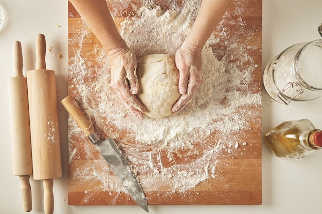 Vista dall'alto sul tavolo bianco con tavola di legno isolata con coltello, due mattarelli, bottiglia di olio d'oliva, barattolo trasparente con farina Le mani della donna tengono l'impasto preparato per pasta o gnocchi