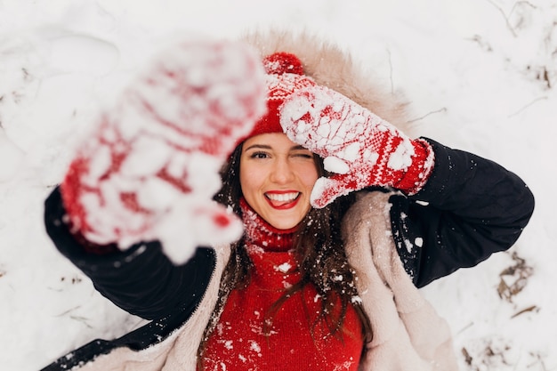 Vista dall'alto sulla giovane donna felice sorridente piuttosto candida in guanti rossi e cappello lavorato a maglia che indossa mantello nero che giace nella neve nel parco, vestiti caldi, divertendosi