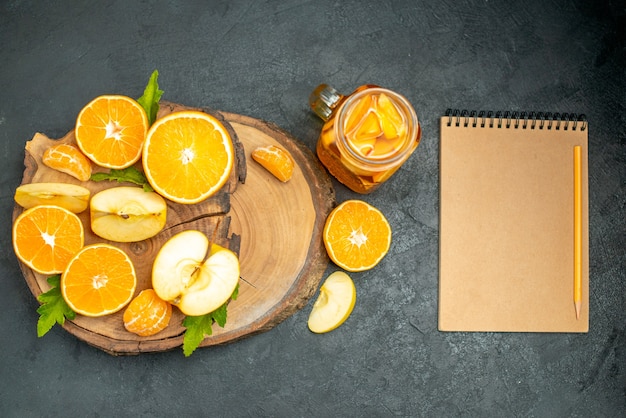 Vista dall'alto tagliare mele e arance su tavola di legno cocktail un quaderno su sfondo scuro