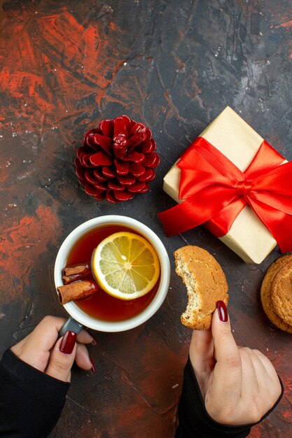 Vista dall'alto tazza di tè aromatizzata al limone e cannella e biscotto in mano femminile regalo di Natale pigna rossa sul tavolo rosso scuro