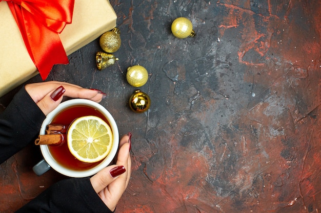 Vista dall'alto tazza di tè aromatizzata al limone e cannella in palline di natale dorate a mano femminile sul tavolo rosso scuro