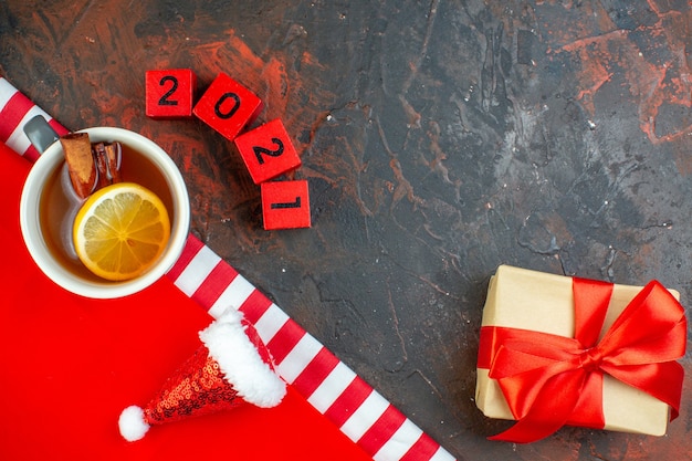 Vista dall'alto tazza di tè aromatizzata al limone e cannella mini cappello di babbo natale su tovaglia rossa regalo in blocchi di legno su tavolo rosso scuro spazio libero